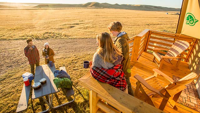 Two young adults on the deck of an oTENTik tent and talking with two other young adults at a picnic table at Grasslands National Park