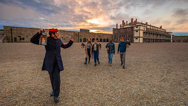 Four visitors and a costumed guide stroll at sunset with lanterns in the courtyard of the Halifax Citadel.