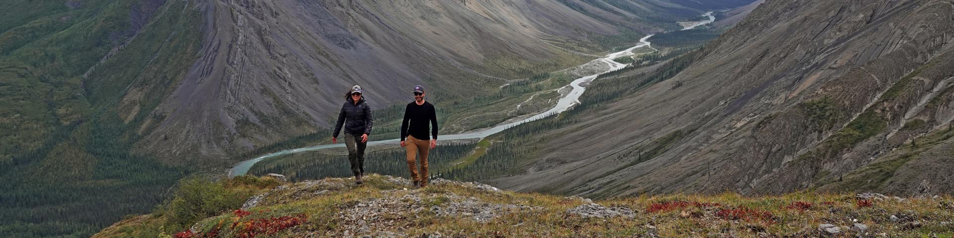 A woman and a man laugh and walk toward the camera, with a view of a river valley and surrounding mountains in the background.
